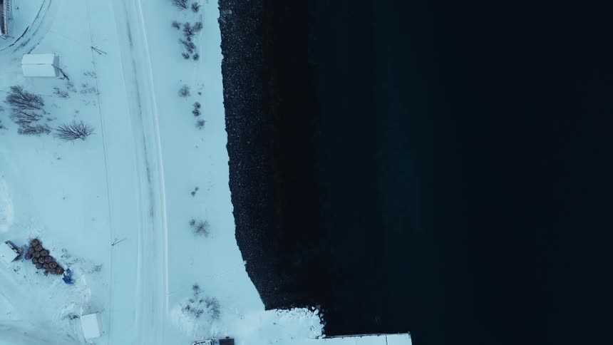 An ascending top-down aerial view of a small, quiet, snow-covered coastal town with a pier next to the dark, cold water