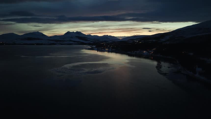 An aerial view shows a tranquil winter fjord at twilight, with snow-covered mountains and a distant village glowing under a dramatic sky