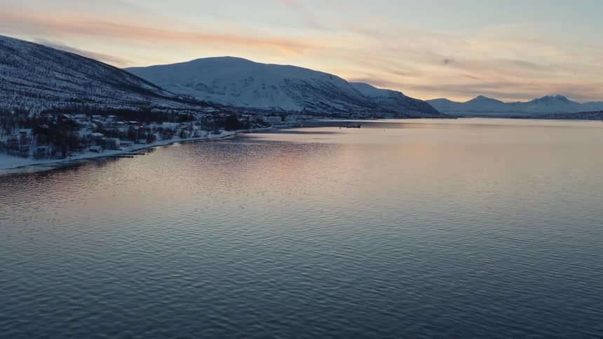 A serene, static aerial view captures the calm waters of a fjord at sunset, with a snow-covered town and mountains along the coastline