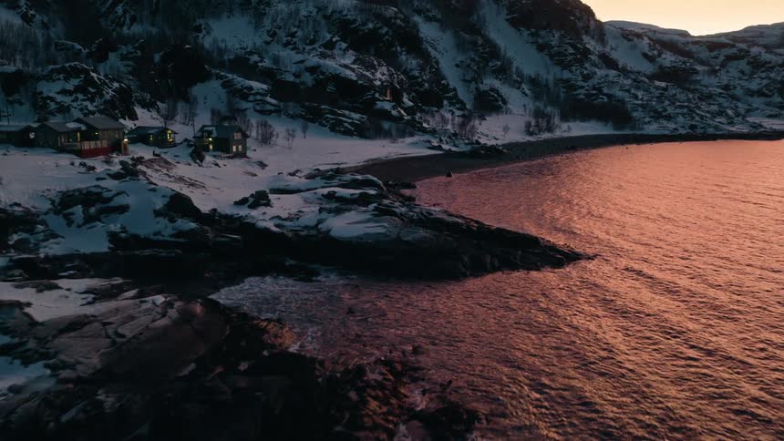 Aerial view of a serene winter coastline with snowy mountains and cozy houses lit up at sunset