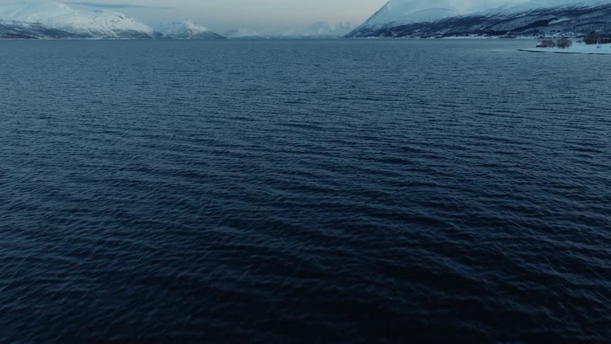 A stunning aerial shot tilts up from the dark, rippling water to reveal a vast, serene fjord surrounded by majestic, snow-covered mountains in Norway during winter