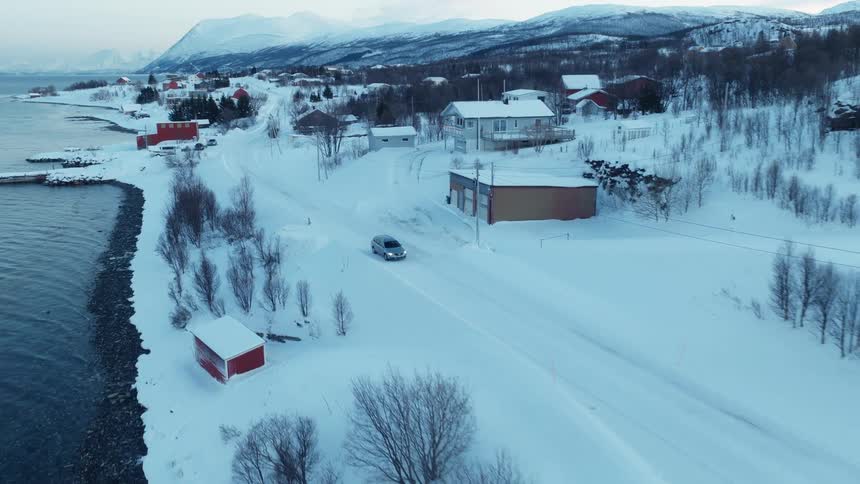 An aerial view follows a car driving along a snow-covered coastal road through a small village in a winter mountain landscape