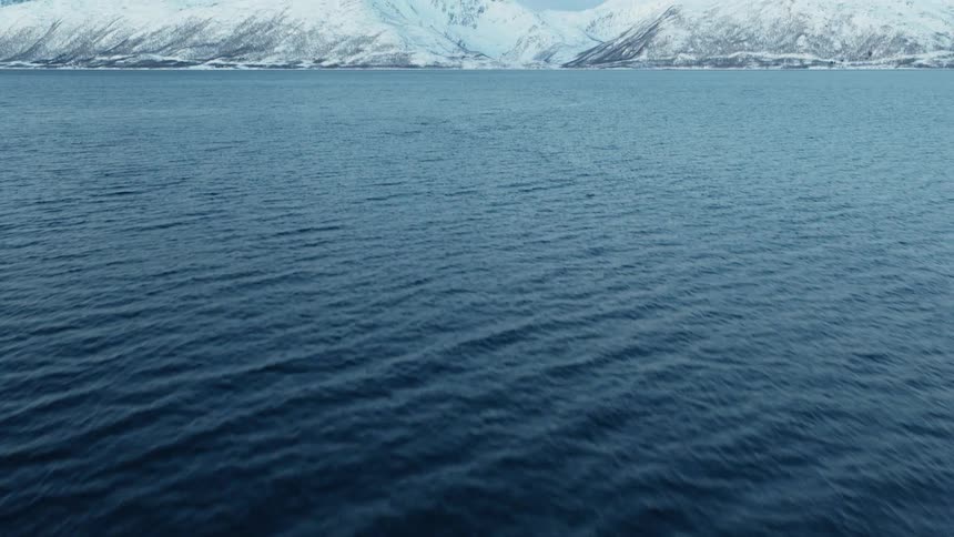 Drone footage slowly tilts up from the rippling surface of a cold, blue fjord to reveal a majestic winter landscape of snow-covered mountains in Norway