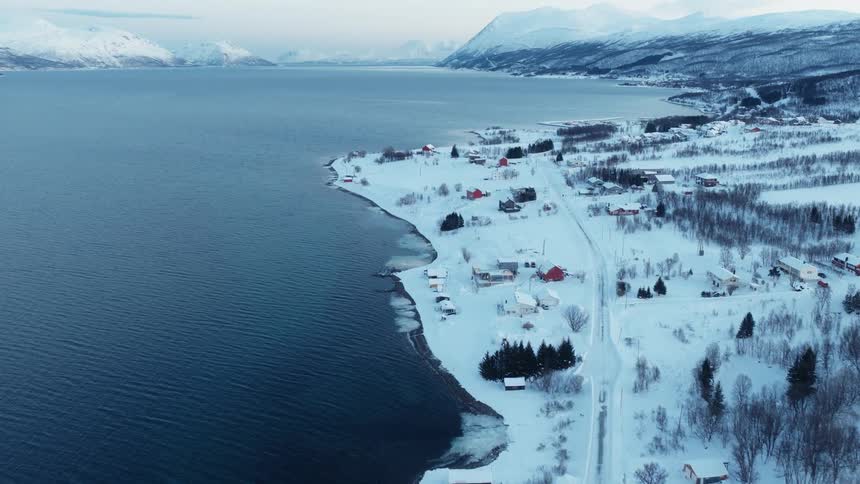 An aerial view tilts up revealing a snow-covered coastal village beside a vast fjord with majestic mountains in the distance