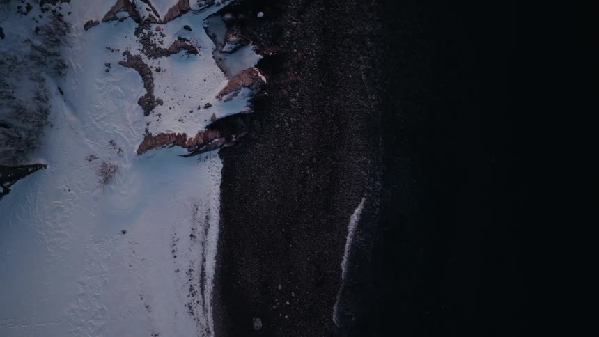 An ascending top-down aerial shot reveals the dramatic contrast between a snow-covered rocky shoreline and the dark, calm waters