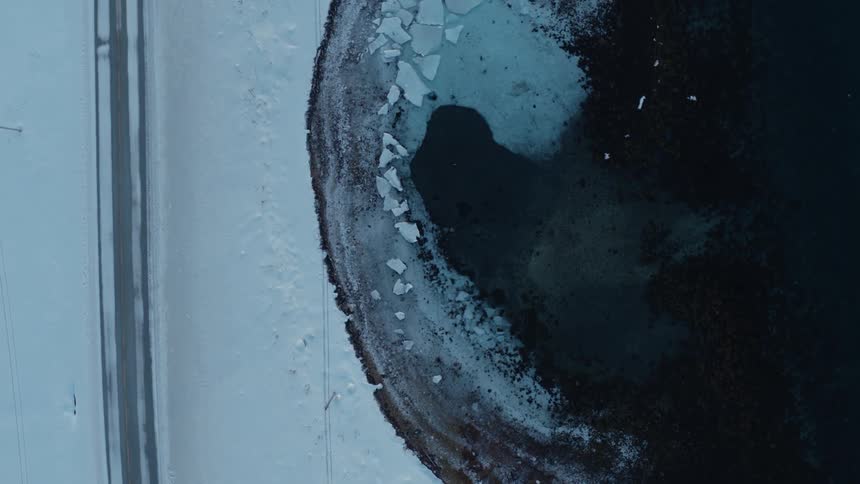 Top-down aerial footage of a car driving on a snowy road alongside a frozen lake shoreline