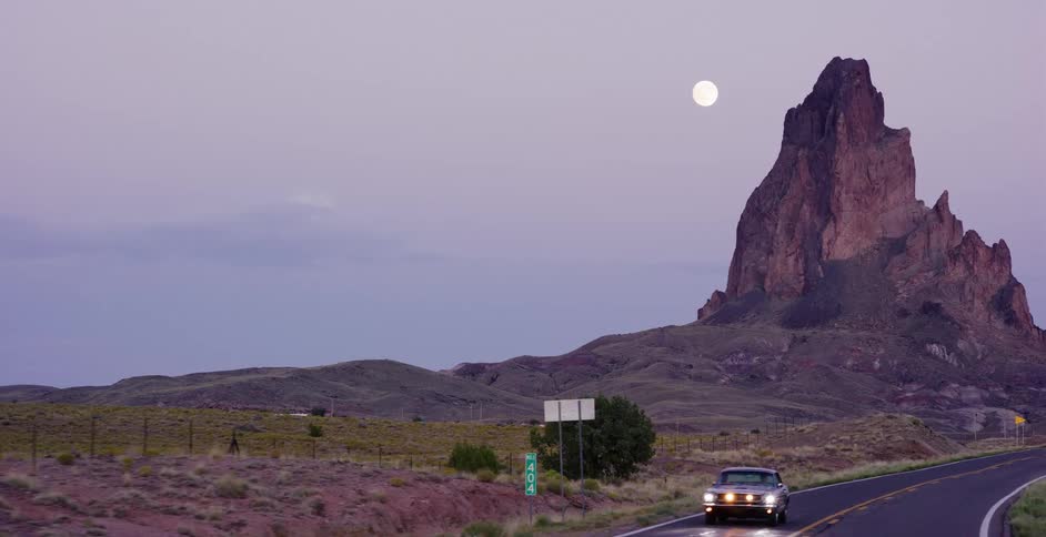 A classic car drives down a desert highway at twilight with a full moon rising over a majestic butte