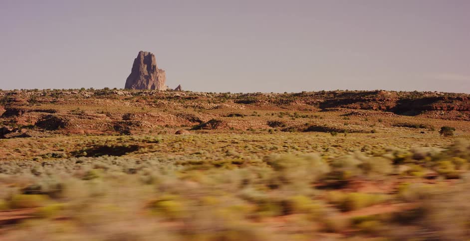 A tracking shot from a moving vehicle captures the vast desert landscape of the American Southwest, featuring a solitary rock formation on the horizon