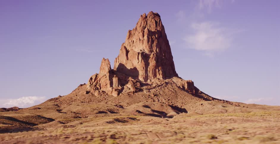 A driving shot passes the iconic Agathla Peak rock formation in the Arizona desert during a warm, golden sunset