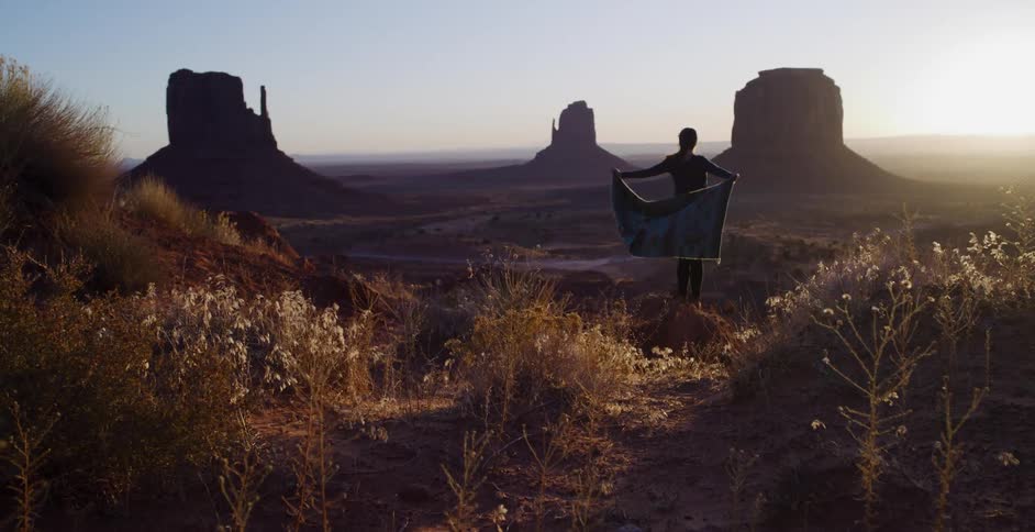 A silhouetted woman stands on a desert ridge holding a tapestry in the breeze with the iconic buttes of Monument Valley in the background at sunset