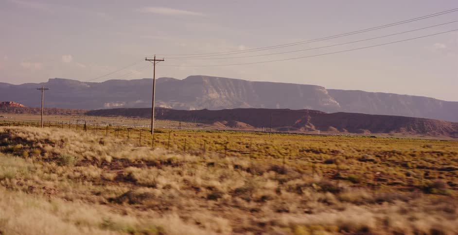 A scenic view from a moving vehicle captures the vast, arid landscape of the American Southwest with distant mesas under a warm sun