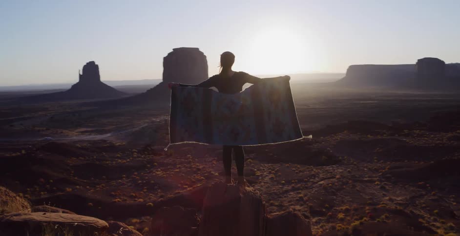 A woman stands on a cliff's edge and unfurls a patterned blanket, silhouetted against the rising sun over Monument Valley