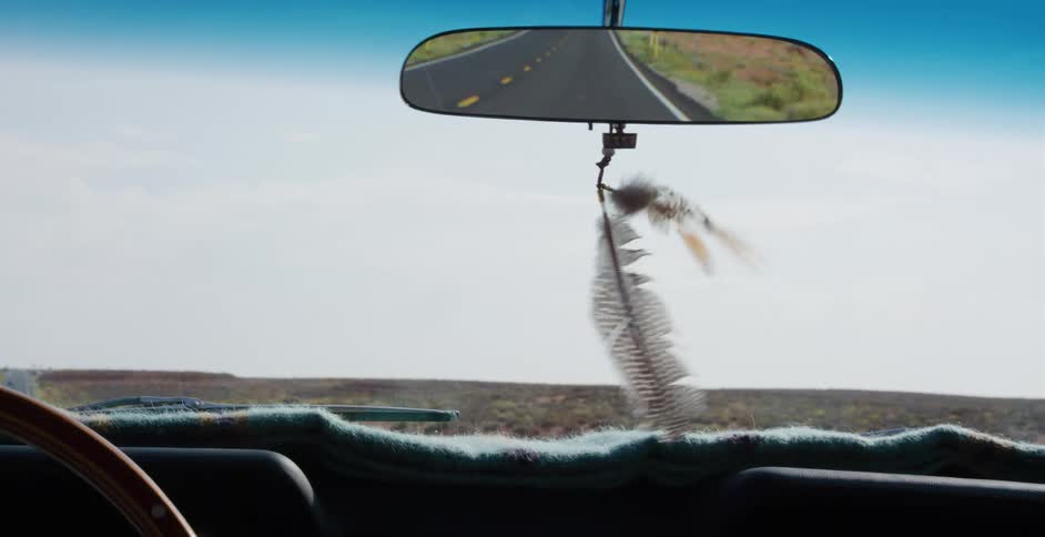 Feathers sway from a rearview mirror as a car drives down an empty desert highway