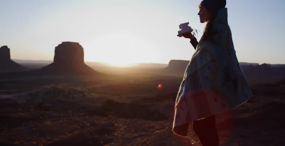 A woman bundled in a blanket stands on a cliff's edge at sunrise, capturing the vast desert landscape with her instant camera