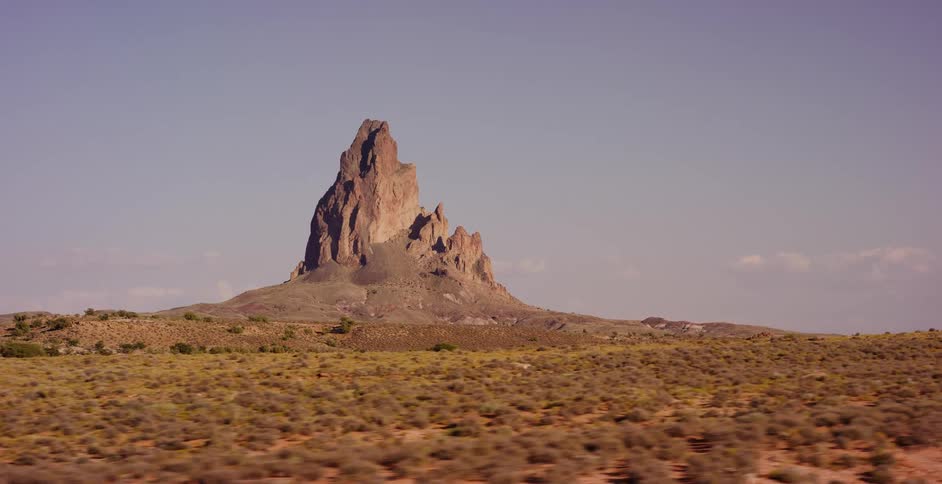 A tracking shot of the majestic Agathla Peak rising from the vast, arid desert landscape in Monument Valley