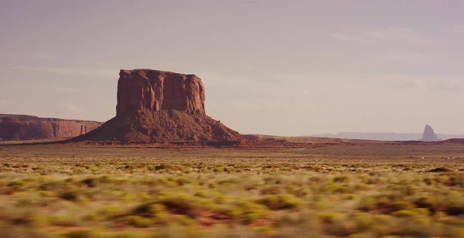 A tracking shot from a moving vehicle captures a majestic red rock butte in the Monument Valley desert