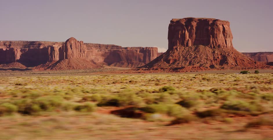 A smooth tracking shot captures the iconic red rock buttes of Monument Valley from a moving vehicle on a sunny day