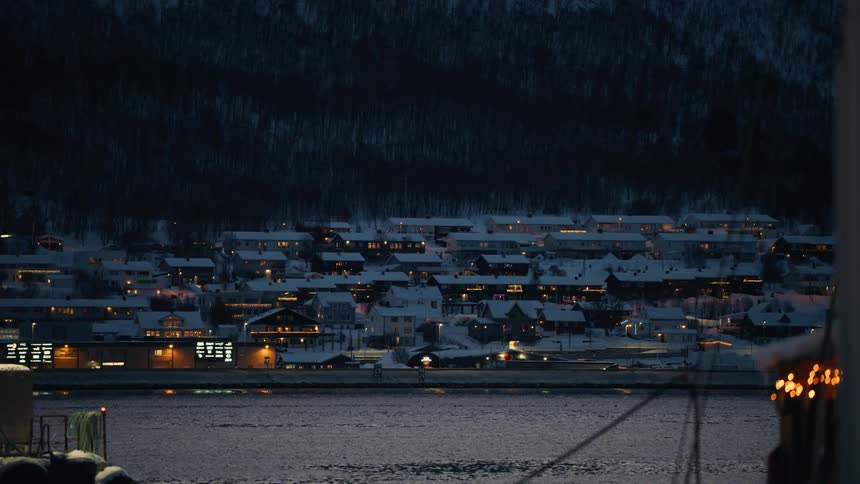 A snowy coastal town in Norway glows with warm lights across the water on a quiet winter night