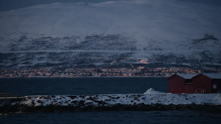 A tranquil view of a small Norwegian town's lights twinkling across a fjord at twilight, set against a backdrop of snow-covered mountains