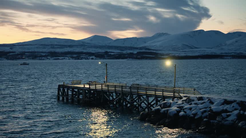 An empty wooden pier with a glowing lamp post sits on a calm fjord at dusk, surrounded by a serene snow-covered mountain landscape