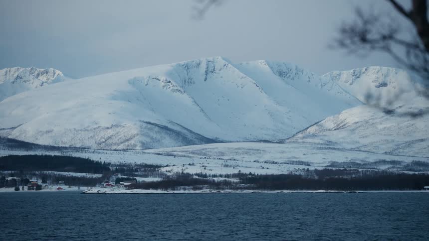 Wide shot of a tranquil fjord in winter with a snow-covered mountain range and a small village on the distant shore