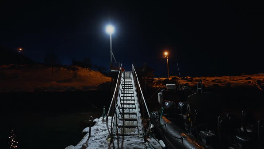 A person walks up a snowy staircase from a floating dock where a rigid inflatable boat is moored on a dark winter night