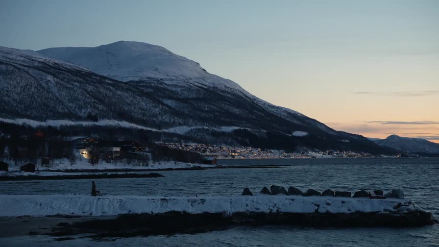 A lone figure walks their dog along a snowy pier at dusk, with the lights of a remote coastal village twinkling against a mountain backdrop