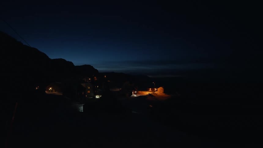 A quiet, remote mountain village glows with warm lights under a dark blue sky during a cold winter night