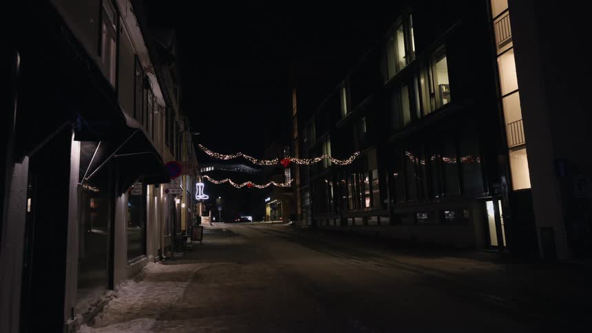 A quiet and empty city street is adorned with festive Christmas lights on a snowy winter night