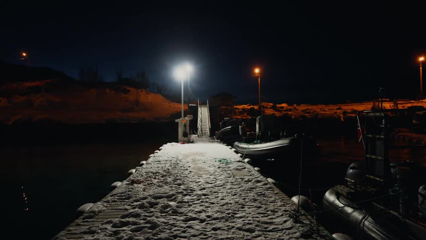 A quiet, snow-covered pier is brightly illuminated by lights on a dark winter night, with inflatable boats moored alongside