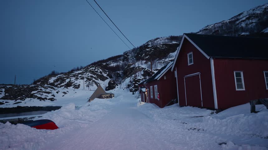 A tranquil scene of a snow-covered road leading past a red wooden house in a remote Scandinavian village at dusk
