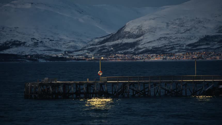 A calm evening view of a lit wooden pier in a fjord with snowy mountains and a distant town in the background
