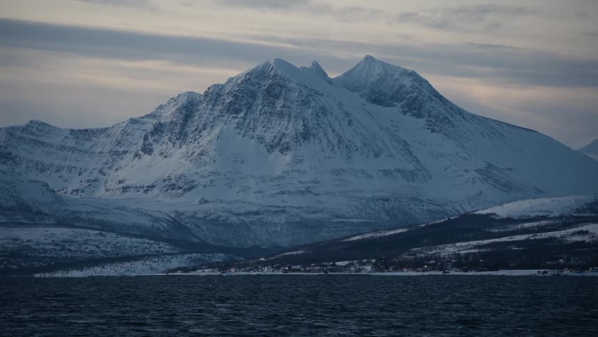 A vast, snow-covered mountain range rises above the cold waters of a fjord where a small village glows at dusk