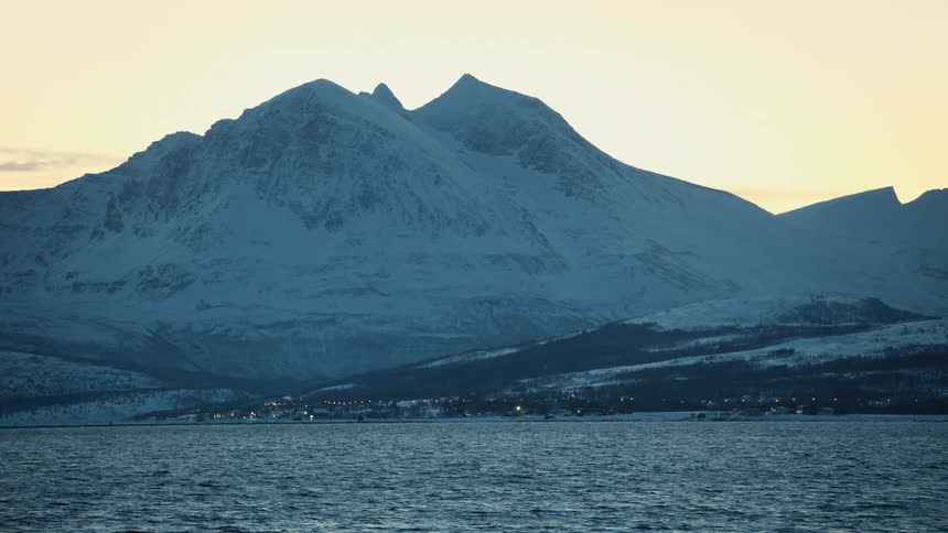 A coastal village with twinkling lights sits at the base of a majestic snow-covered mountain range on the edge of the sea at dusk