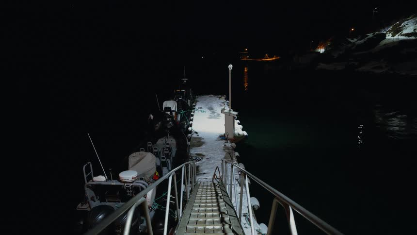 A ramp leads down to a snowy dock where two boats are moored in the calm, dark water on a quiet winter night