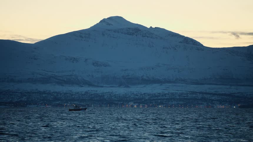 A lone boat sails across a vast, chilly body of water at twilight with a majestic snow-covered mountain and distant city lights in the background