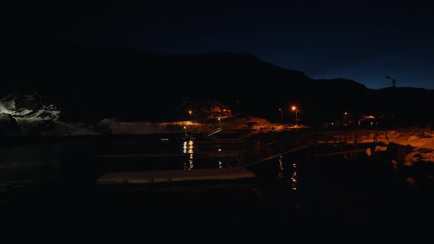 A tranquil snowy shoreline at night with a small pier and streetlights reflecting on the calm, dark water