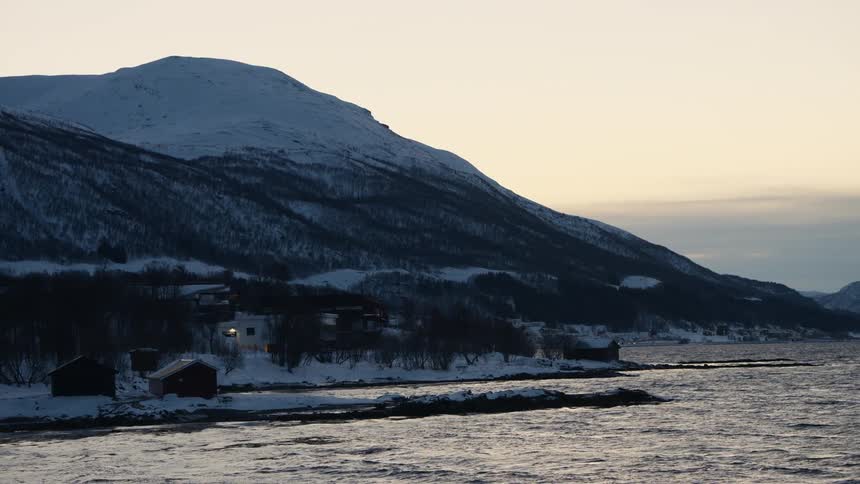 A tranquil winter scene of a snowy Norwegian coastline with cozy houses at the base of a large mountain at dusk