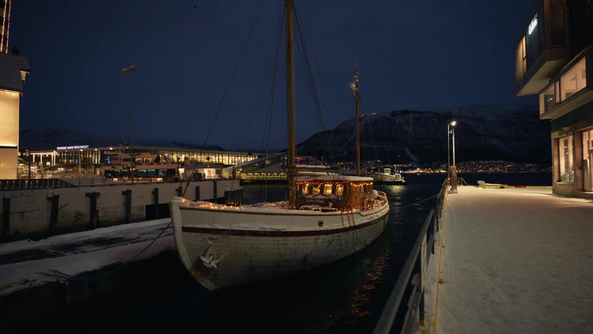 A classic wooden sailboat adorned with warm festive lights is docked in a snowy harbor at night, with city lights and mountains in the background