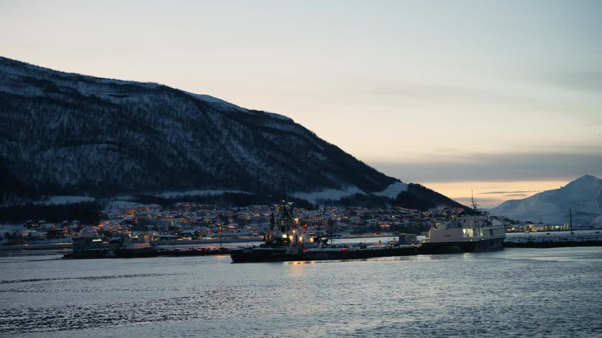 A tranquil harbor with boats is illuminated by town lights at the base of a snowy mountain during a winter twilight