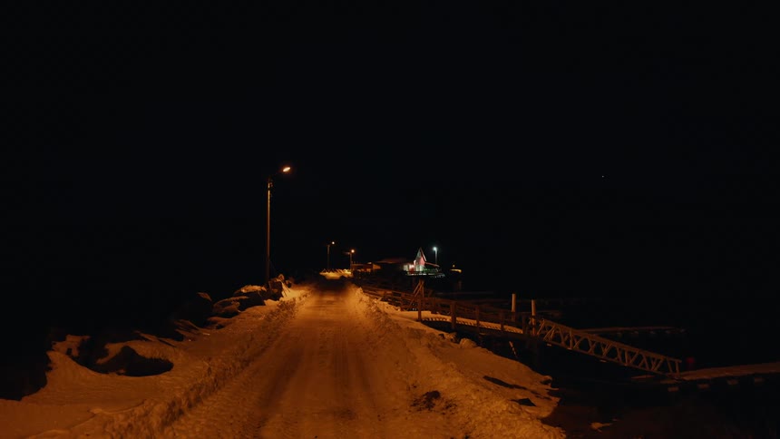A snow-covered road leads to a brightly lit building on a pier during a still, dark winter night