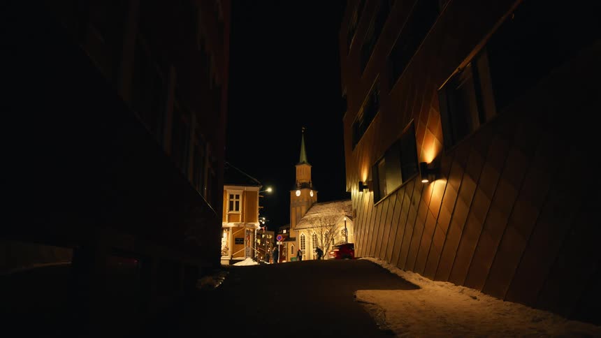 An atmospheric night view from a dark alley towards a brightly lit church in a snowy European town