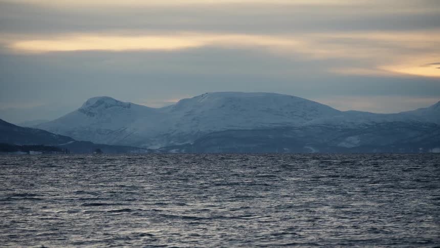 A tranquil winter scene of a vast body of water with snow-capped mountains on the horizon under a soft, cloudy sunset sky