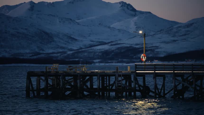 A lonely illuminated pier juts out into a calm fjord at dusk, with snowy mountains in the background