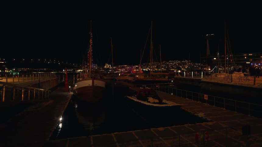 Sailboats decorated with holiday lights rest in a calm, snowy harbor at night with the twinkling lights of a city in the background