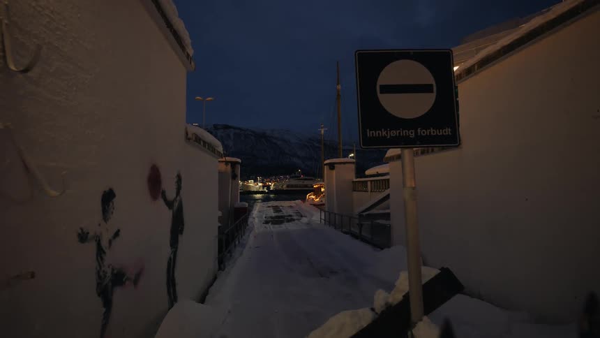 A snowy back alley with street art and a no entry sign leads to a harbor as a ferry passes at night in Norway