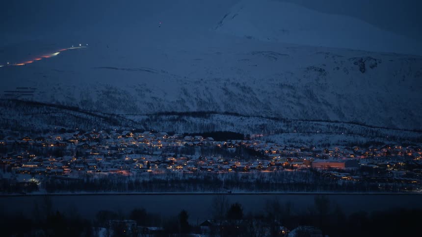 A tranquil view of a small snowy village nestled at the foot of a vast mountain, with warm lights glowing during the cold blue hour