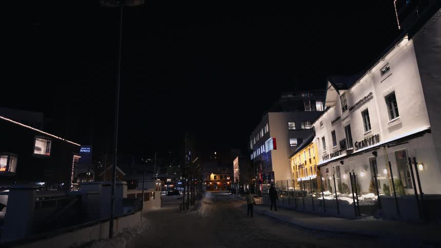 A tranquil, snow-covered street in a small town is warmly illuminated by building lights on a dark winter night