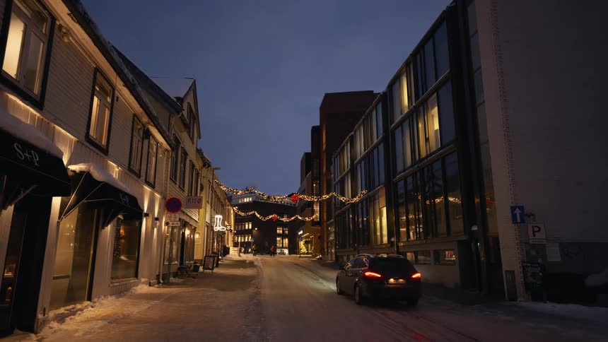 A car drives slowly down a quiet, snow-covered city street decorated with Christmas lights at dusk