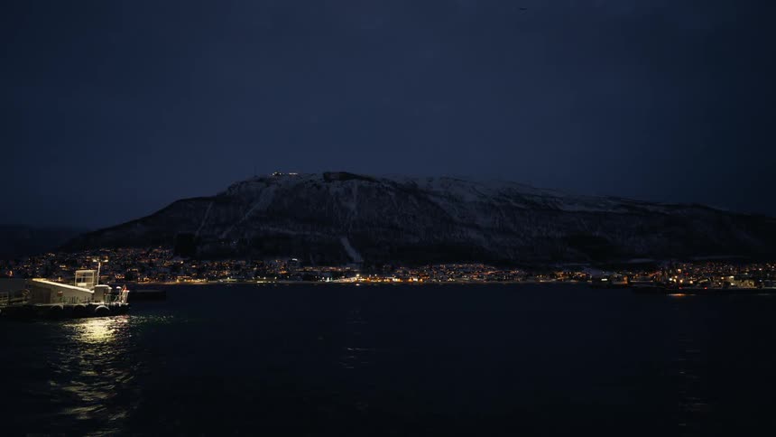 A picturesque northern city glimmers across the dark water, nestled against a vast, snow-covered mountain at twilight